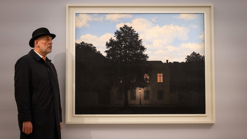 A man in a black coat and hat stands next to Rene Magritte's L'empire des lumieres,1961.