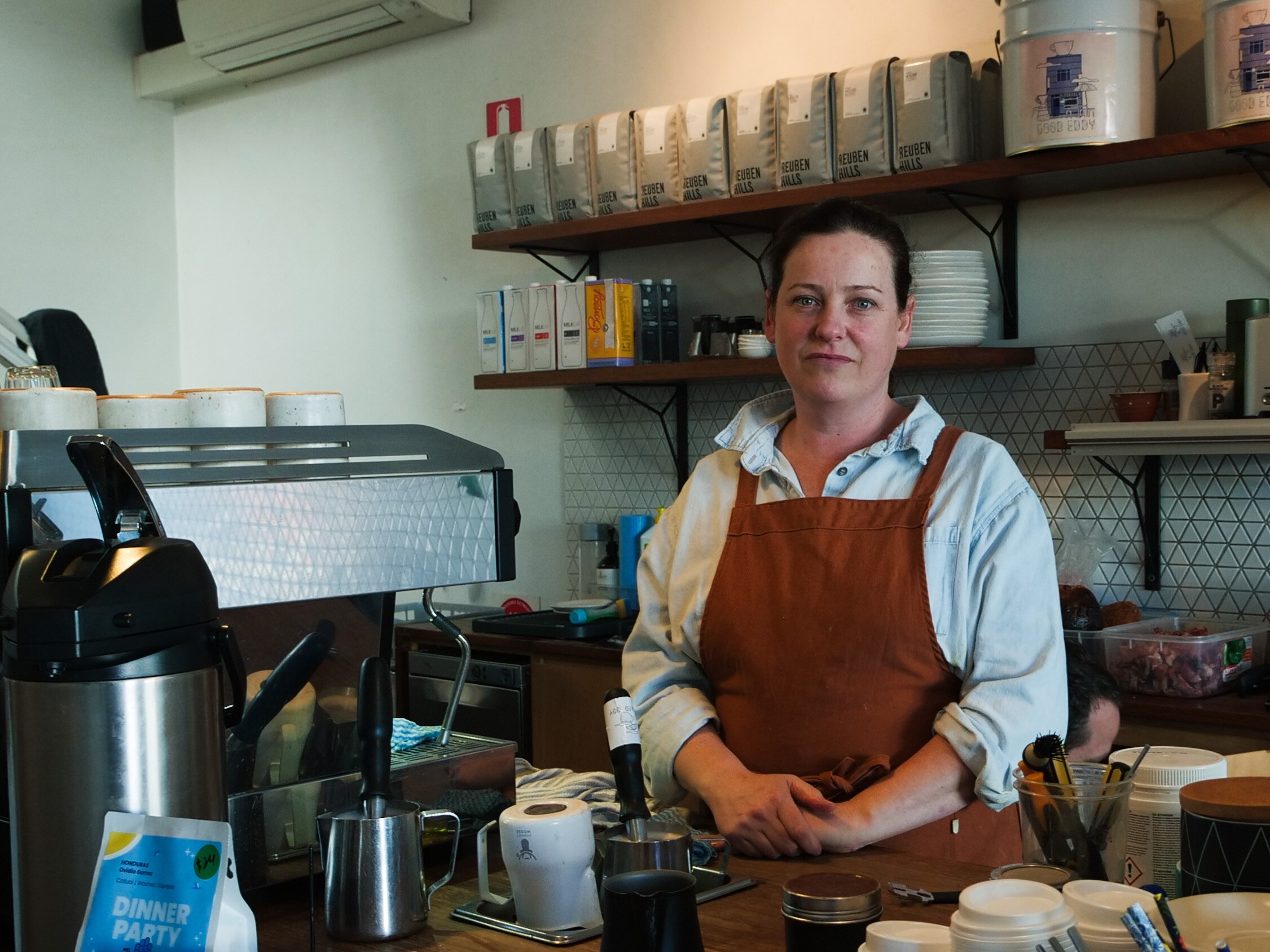 A woman in a brown apron stands frowning behind a counter in a cafe with a coffee machine.