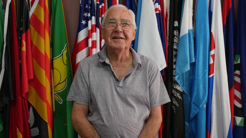 82-year-old Ron Strachan standing in front of wall of colourful flags.