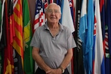 82-year-old Ron Strachan standing in front of wall of colourful flags.