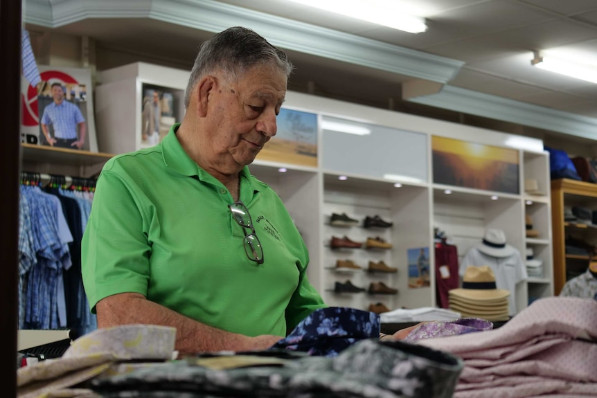 An elderly man holds a shirt in his hands, in a men's clothing store.