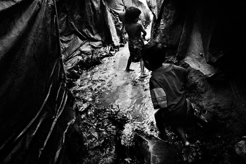 Two Rohingya children play in a flooded walkway between tents.
