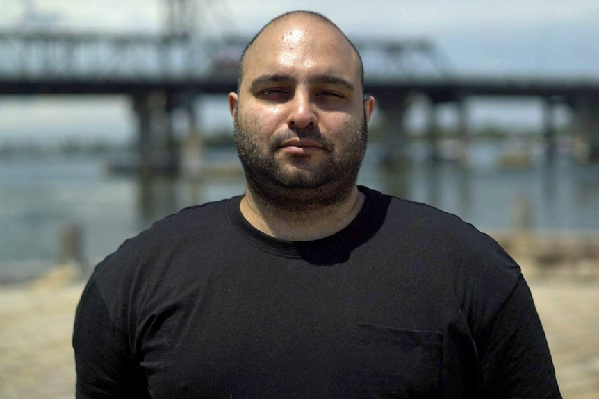 Man in black t-shirt stands in front of a bridge.