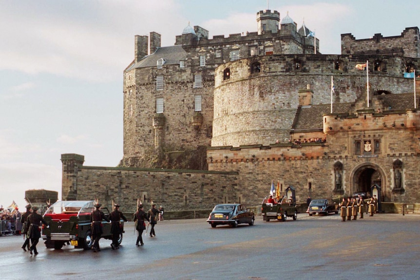 A Range Rover flanked by Royal Archers drives up towards a giant castle.