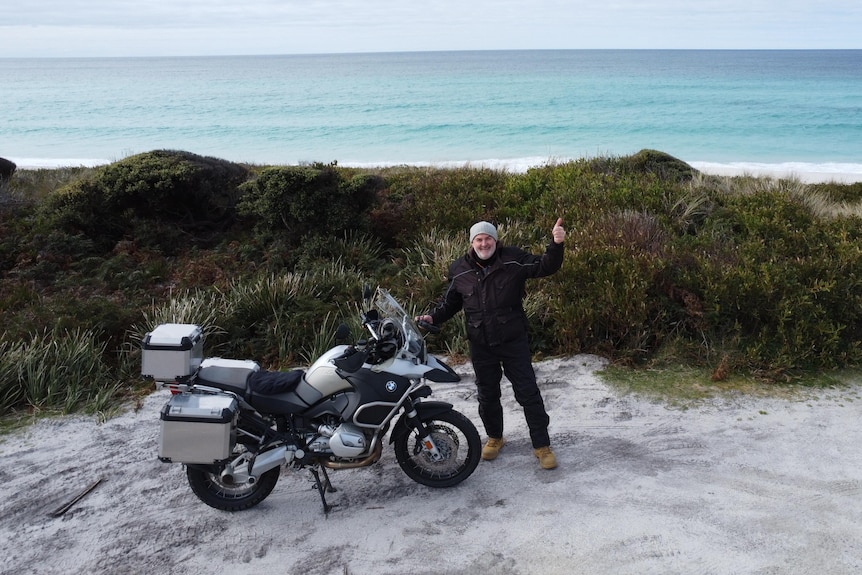 A man smiles and gives a thumbs-up as he stands next to a motorbike. Behind him stretches a beautiful beach.