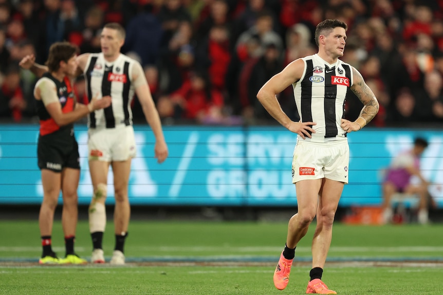 A Collingwood player stands with hands on hips at the MCG as Magpies and Bombers players shake hands in the background.