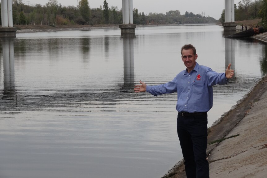 A man in a blue long-sleeved shirt stands next to a wide canal with his arms outstretched.