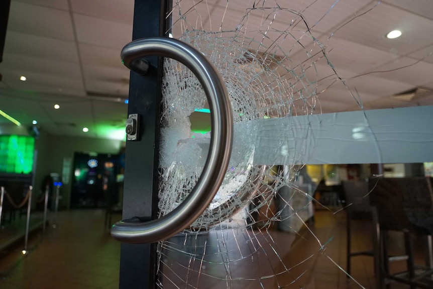 A glass door is seen with cracks running through it, with a hole in the middle. The green light of the bar is visible behind it.