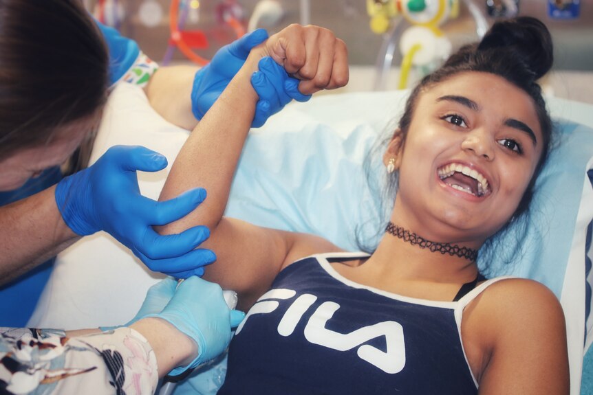 A girl smiles while laying on a hospital bed getting an injection in her arm.