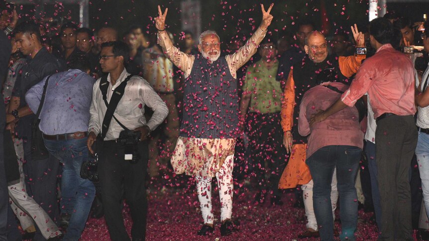 A man in Indian garb holds up his hands in victory signs while walking through a cloud of rose petals