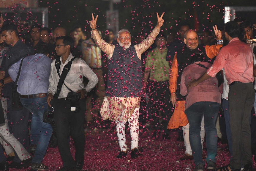 A man in Indian garb holds up his hands in victory signs while walking through a cloud of rose petals