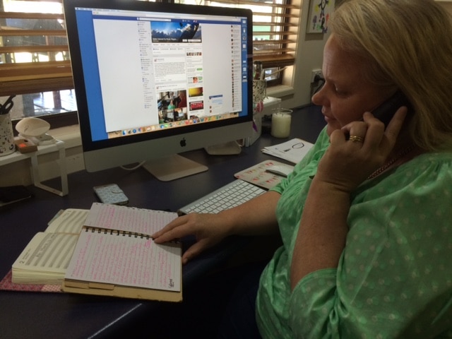 A woman is working at her computer speaking on the phone and looking at a notebook she seems really busy