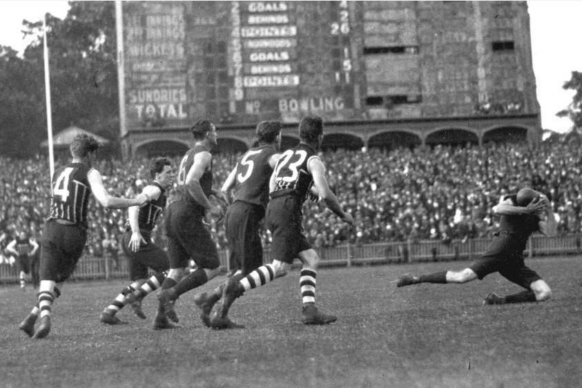 A group of Australian rules footballers watch as one player takes a mark at Adelaide Oval with the old scoreboard in background.