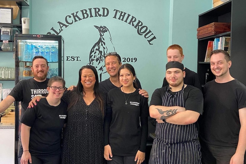 A group of eight people wearing black work shirts stand in a cafe in front of a green wall with a black drawing of a bird.