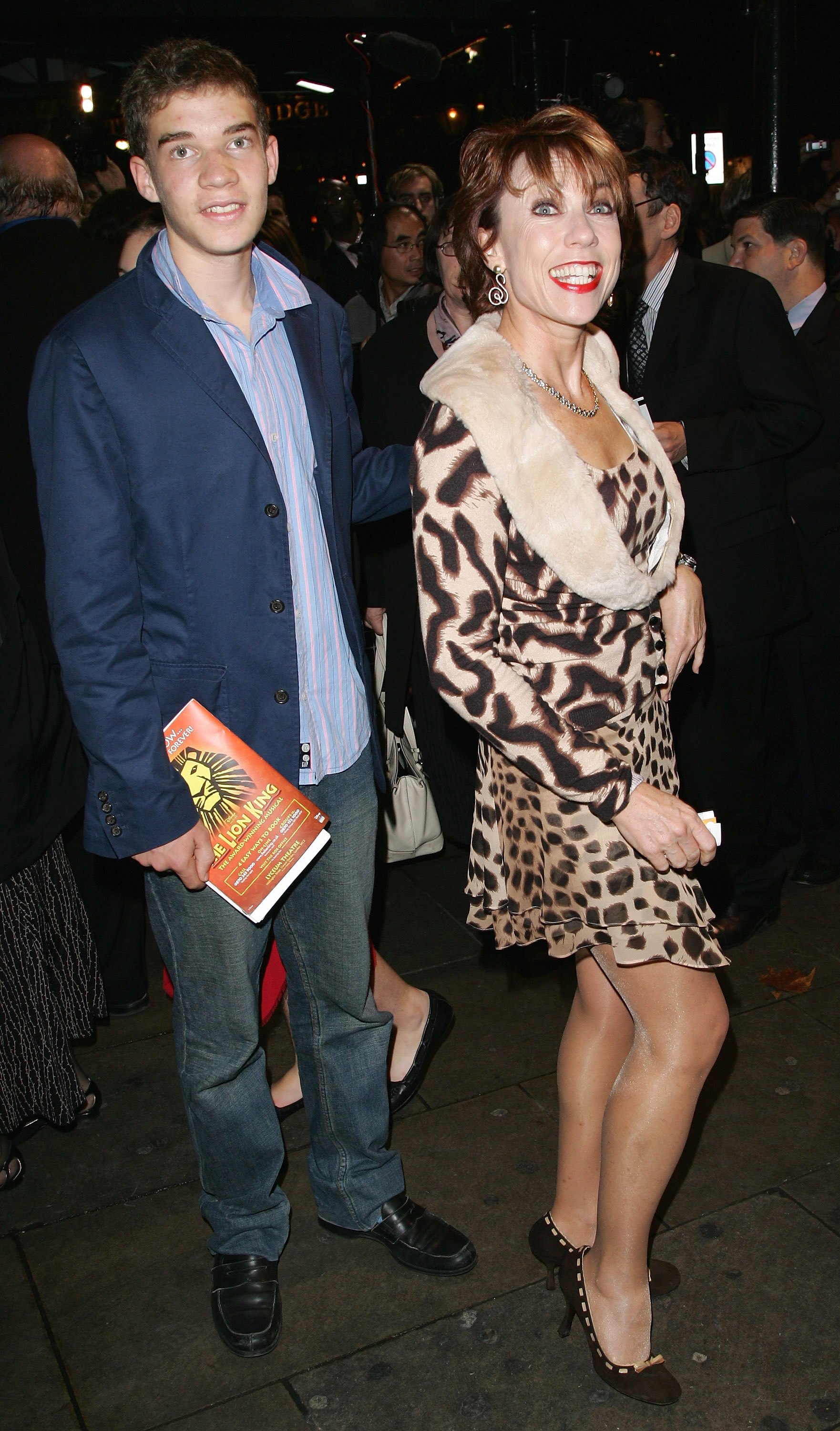 Woman with short brown hair wears cheetah print short dress on red carpet next to her teenage son who holds a theatre playbook