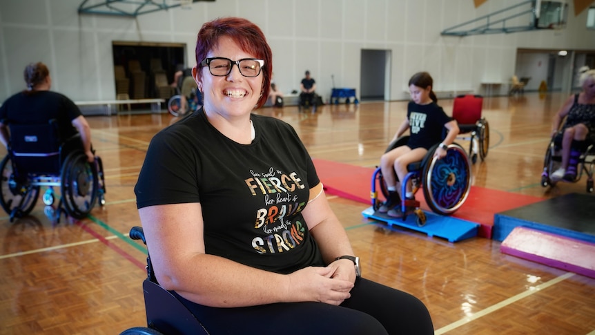 A woman named Melinda Rea smiles while sitting in a wheelchair at a gymnasium.
