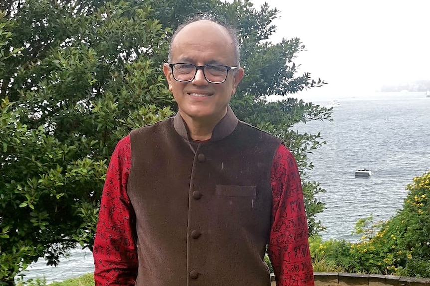 An Indian man in red shirt and brown vest with sea and greenery in the background.