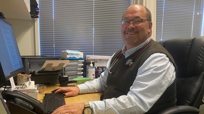 A man wearing a vest and glasses sits at his office desk in front of a computer.