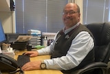 A man wearing a vest and glasses sits at his office desk in front of a computer.