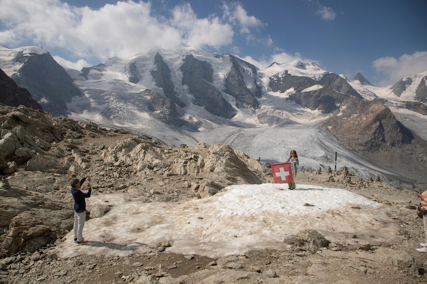 A woman poses for a person taking a photo with a Swiss flag with the alps in the background. 