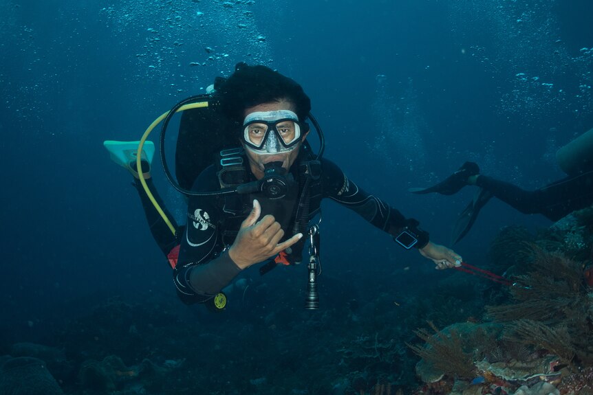 A man in scuba gear gives a thumbs up underwater with a coral reef behind him