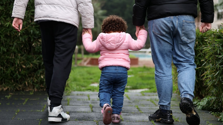A toddler walking and holding her parents hands