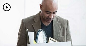 A man wearing a jacket and jumper holds a box filled with office paraphernalia, while looking down at it with sombre expression.