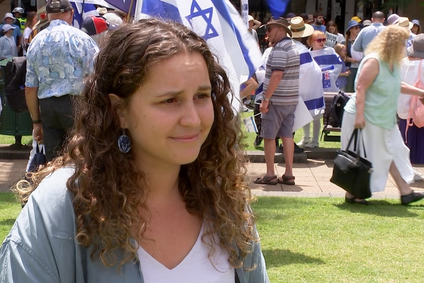 a woman standing in a park at a pro-israel rally, flags can be seen in the background
