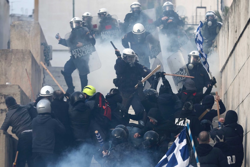 Protesters and riot police both clad in black clash with each other up steps—protestors attacking police with Greek flagpoles