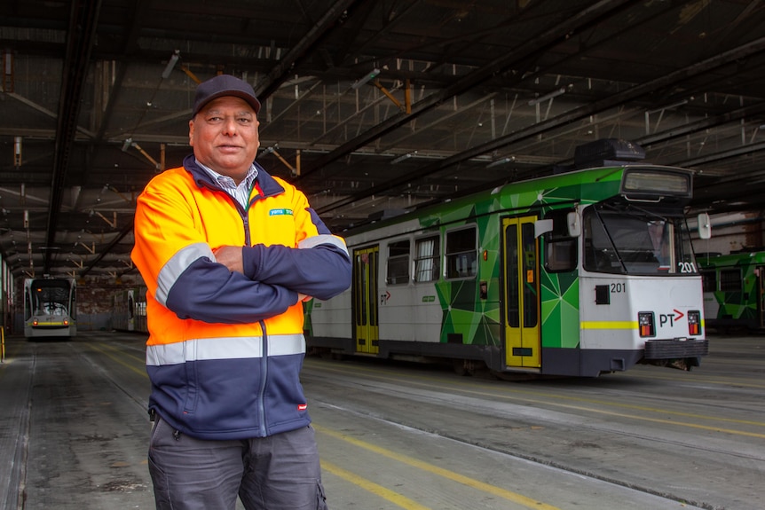 A middle age man with black cap, high vis jacket standing in a big shed with trams in it