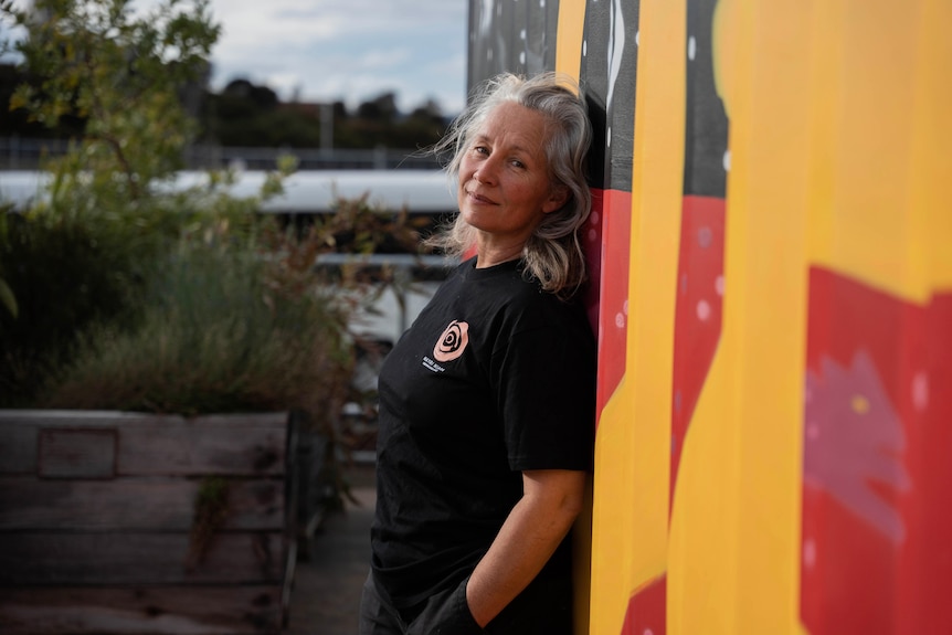 Woman stands with her back against a wall painted in the colours of the Aboriginal flag