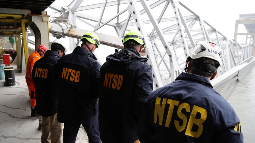 Four men with their backs turned wearing navy coloured jackets with the letters NTSB in yellow written, looking over at water