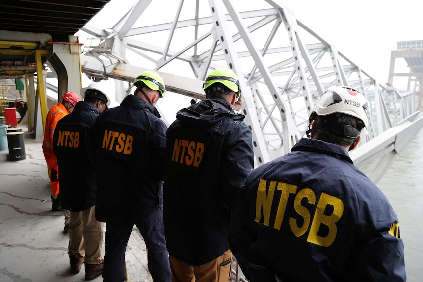 Four men with their backs turned wearing navy coloured jackets with the letters NTSB in yellow written, looking over at water