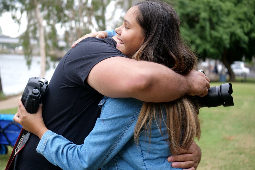 A man and a teenage girl hugging, both holding cameras, grass and river in background.