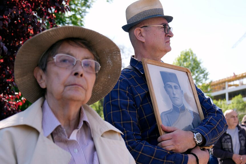 An elderly woman and man attend a march, with the man holding a picture of a deceased relative.