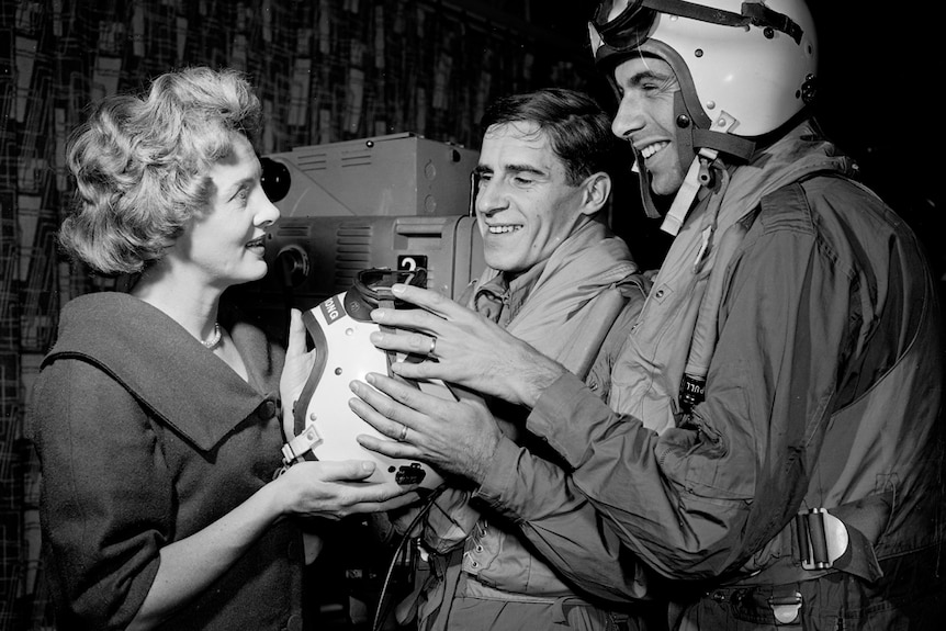 Black and white photo of two men and a woman smiling holding an a helmet. TV cameras are visible behind them.