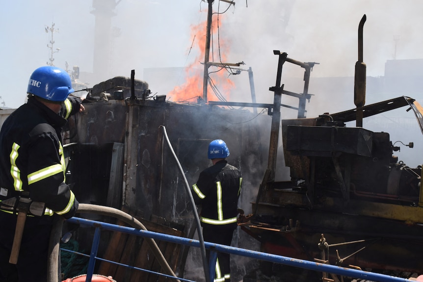 Dos bomberos sostienen una manguera para rociar agua en un incendio en el lado de babor.