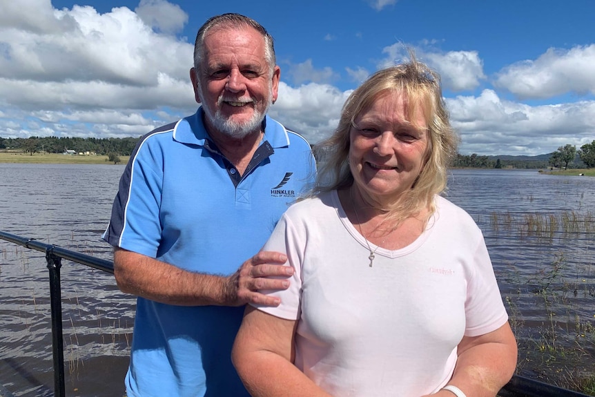 A man and a woman stand, smiling, in front of a full dam.