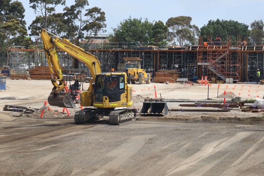 A yellow excavator works in front of the structural skeleton of a building