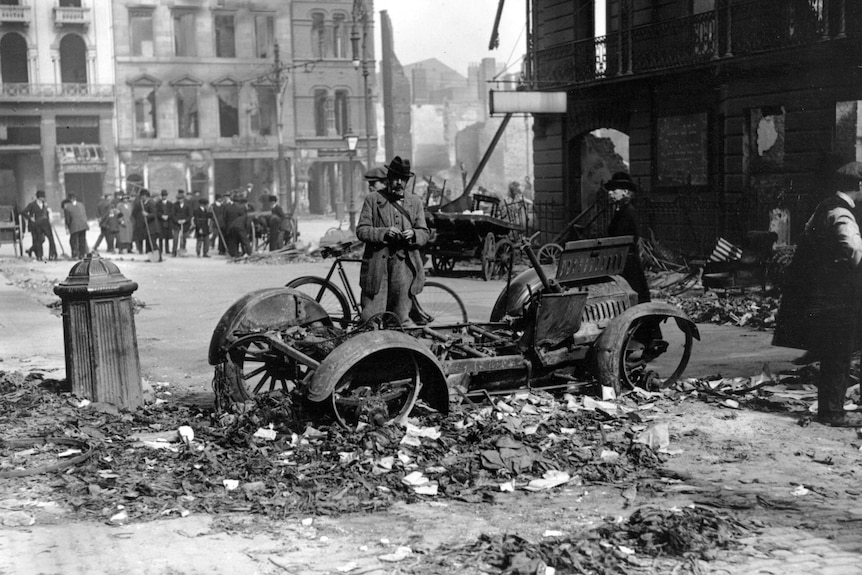 Black and white image of a man standing beside a burnt out car