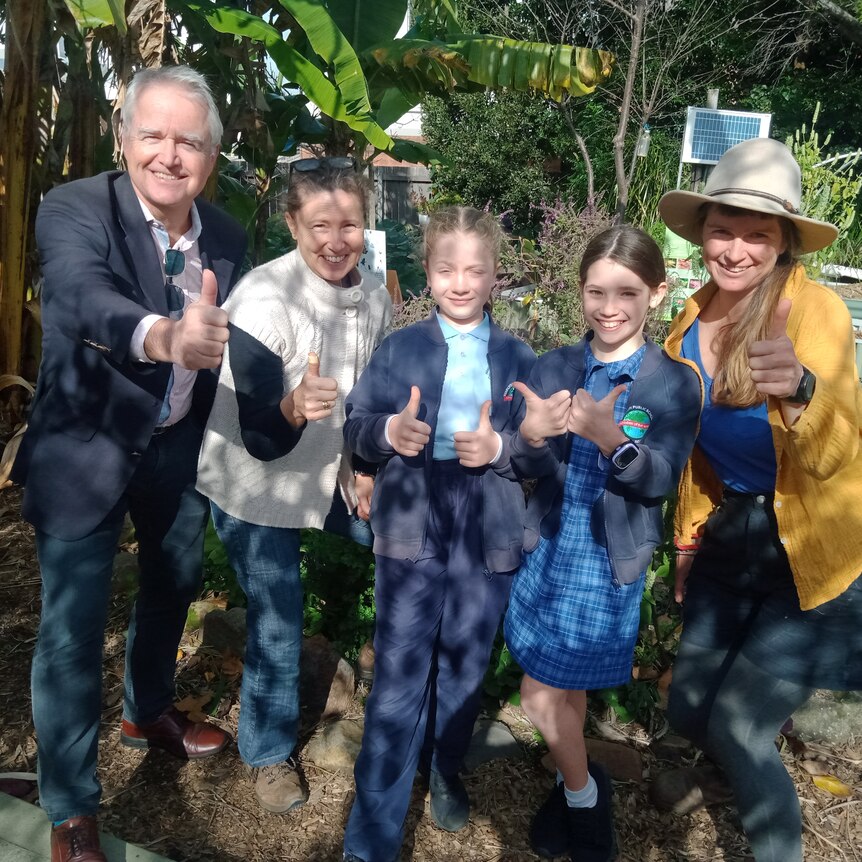 a group of people standing near a garden, they're looking at the camera smiling and giving a thumbs up.