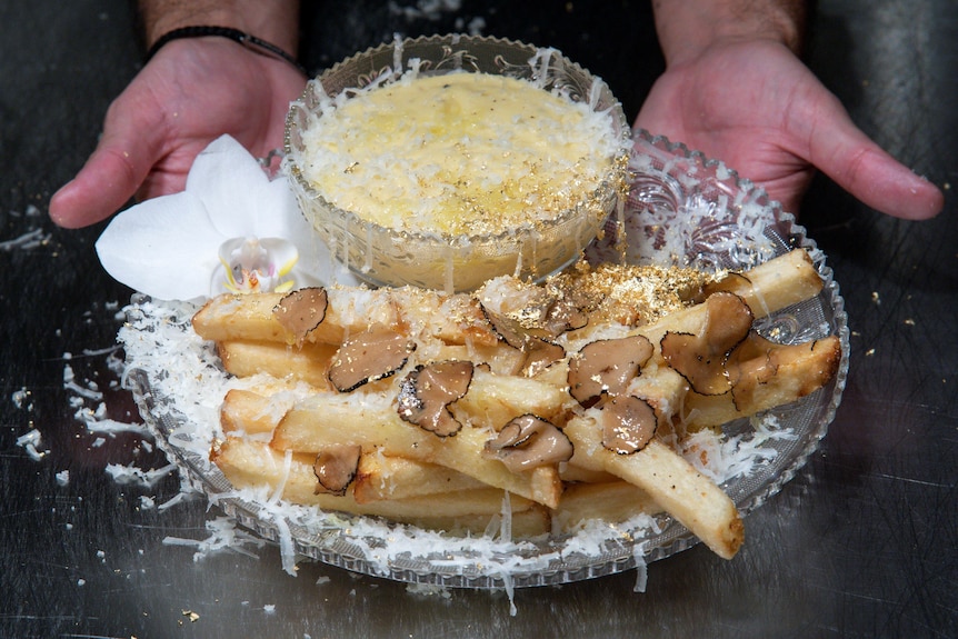 Fries, dusted in gold, grated cheese and covered in thinly sliced pieces of truffles are plated next to a bowl of cheese.