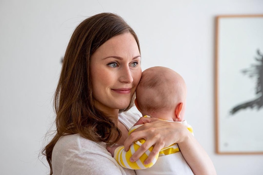 Sanna Marin, a young woman in plain white T-shirt, hugs a baby to her chest. The baby is in a yellow and white striped romper