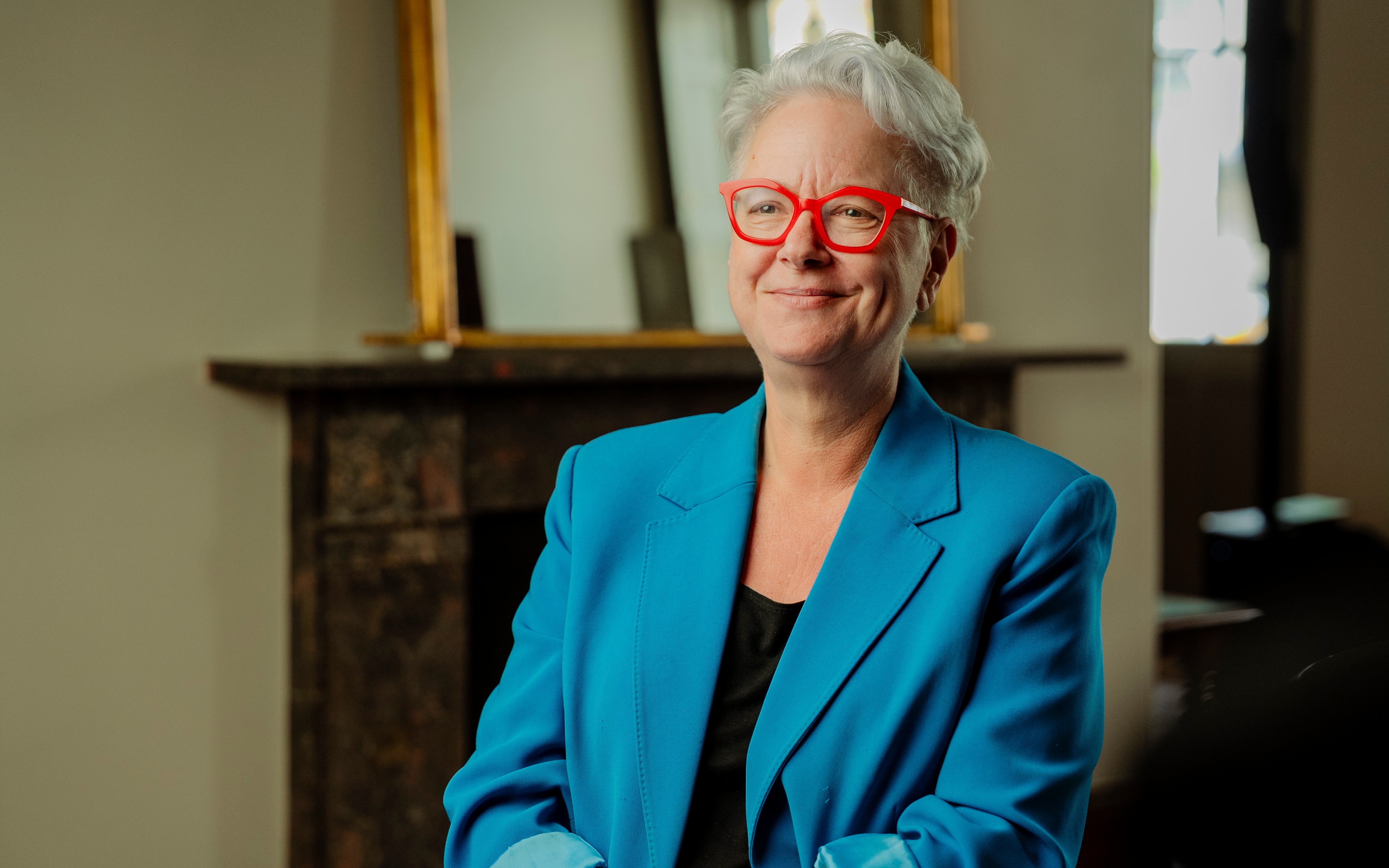 Ms Sharpe sitting in a room on a chair smiling while wearing a blue blazer.