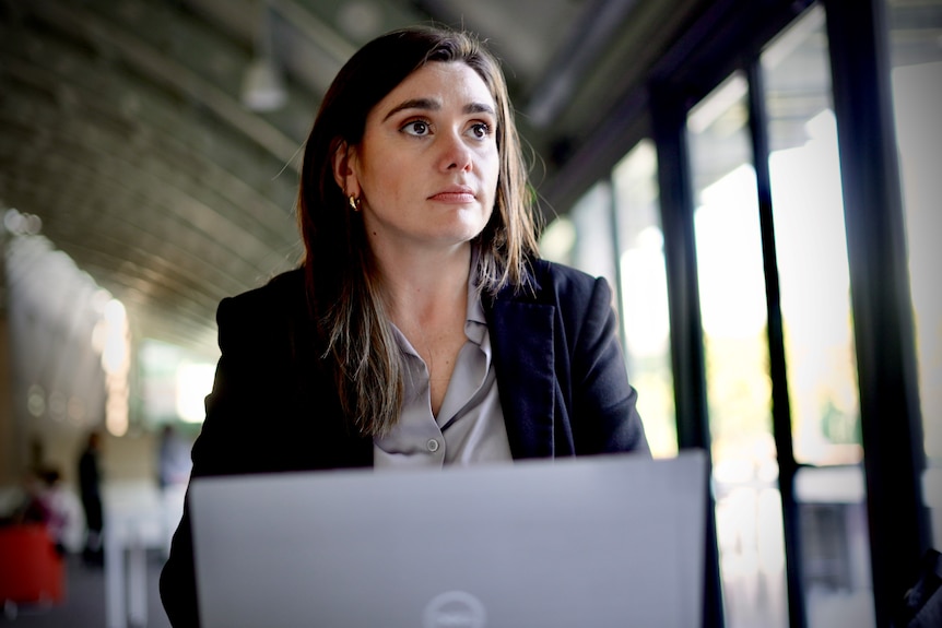 A woman with dark hair and a dark blazer sits at a table with a laptop looking into the distance.