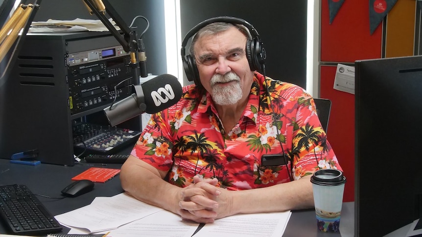 Ray Dickson sits behind the radio broadcasting desk with a microphone, coffee, and a bright Hawaiian shirt