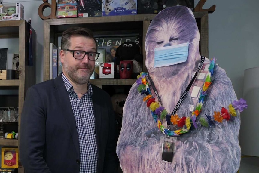 A man in front of a bookcase and next to a cut-out of Chewbacca wearing a face mask