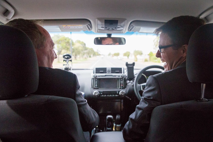 Barnaby Joyce, in the passenger's seat, smiles across at David Littleproud in the driver's seat. They are both wearing suits.