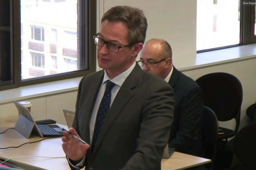 A man wearing a suit stands and speaks in a court room