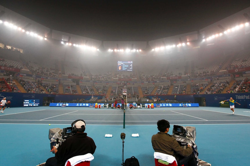 Players play a rally on a tennis court as smog lingers above near the flood lights